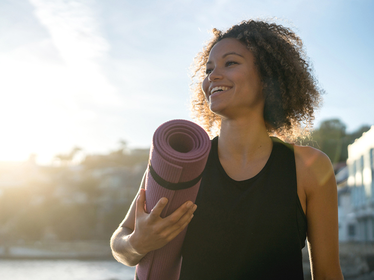 A woman smiles and holds a purple yoga mat as the sun rises just outside the frame.