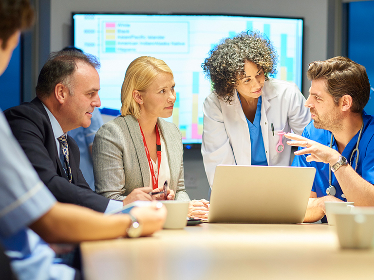 A doctor explains something on a laptop screen to another doctor, a woman with a lanyard, a man with a tie, and another person gathered around the table as a presentation takes place in the background.