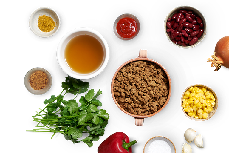 A handled terra cotta bowl of Quorn Mince surrounded by smaller bowls of spices, sauces, beans, and an onion and a bunch of coriander.