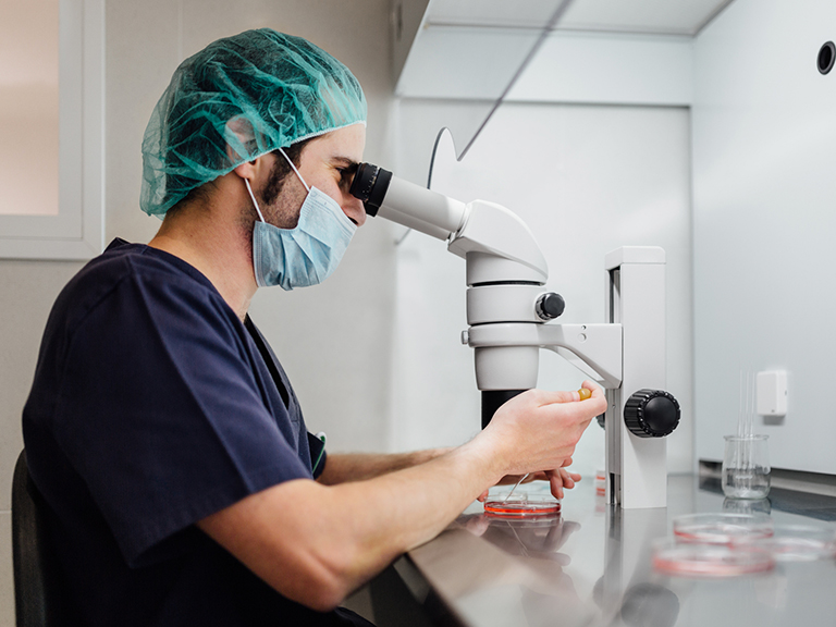 A researcher in a lab looks into a microscope and uses a pipette on the dish under the microscope.